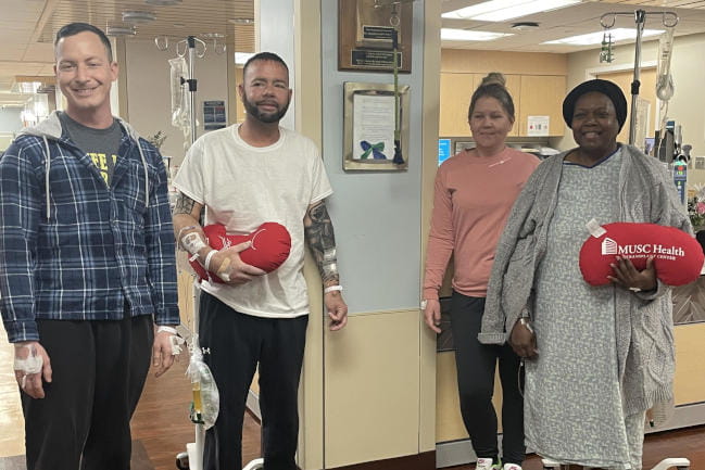 Four people stand in a hospital waiting room, some with IVs attached. They are holding plush pillows shaped like kidneys with the 168极速赛车在线开奖网站 Health logo printed on them.