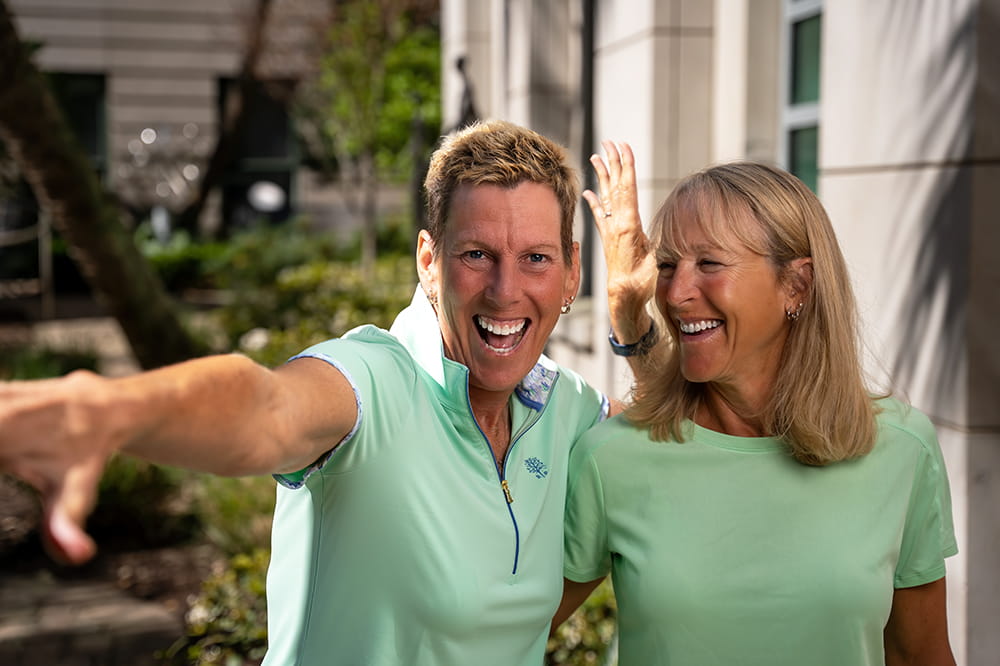 a woman points off camera in a goofy manner while another woman looks at her and laughs