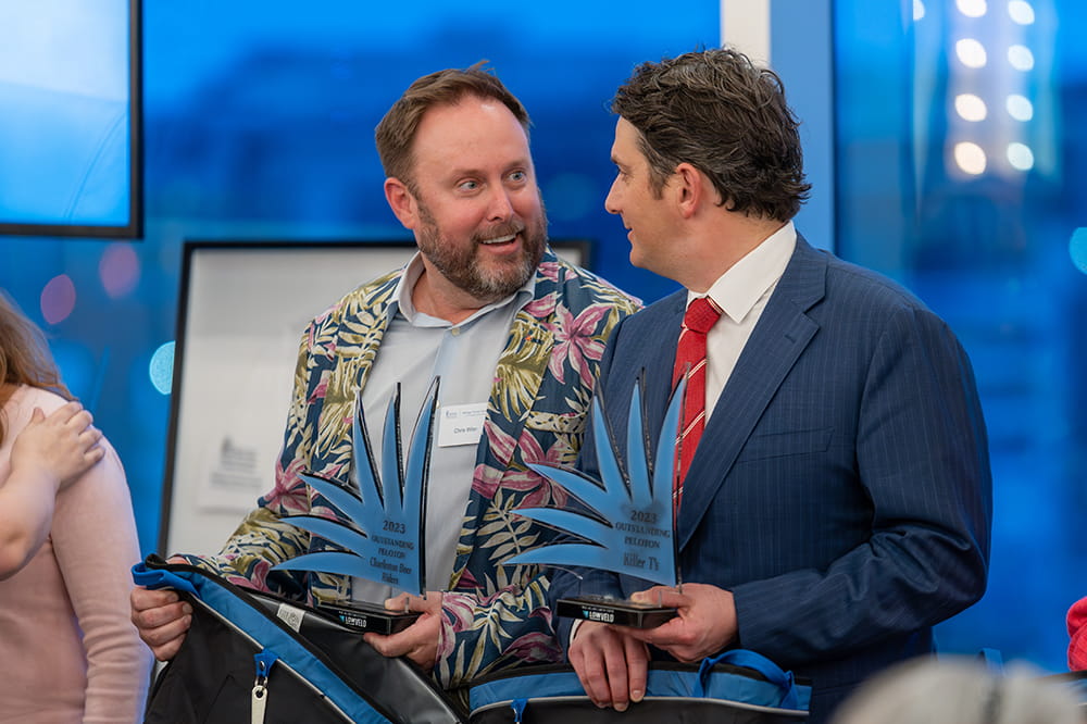 a man in a loud floral sports coat and a man in a blue suit and red tie sit next to each other, grinning and holding acrylic trophy plaques in an abstract shape of palm leaves