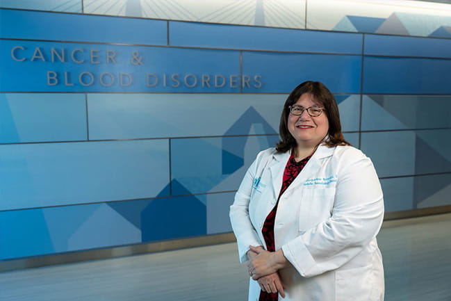 A Caucasian woman in a lab coat stands in front of a sign reading Cancer and Blood Disorders.